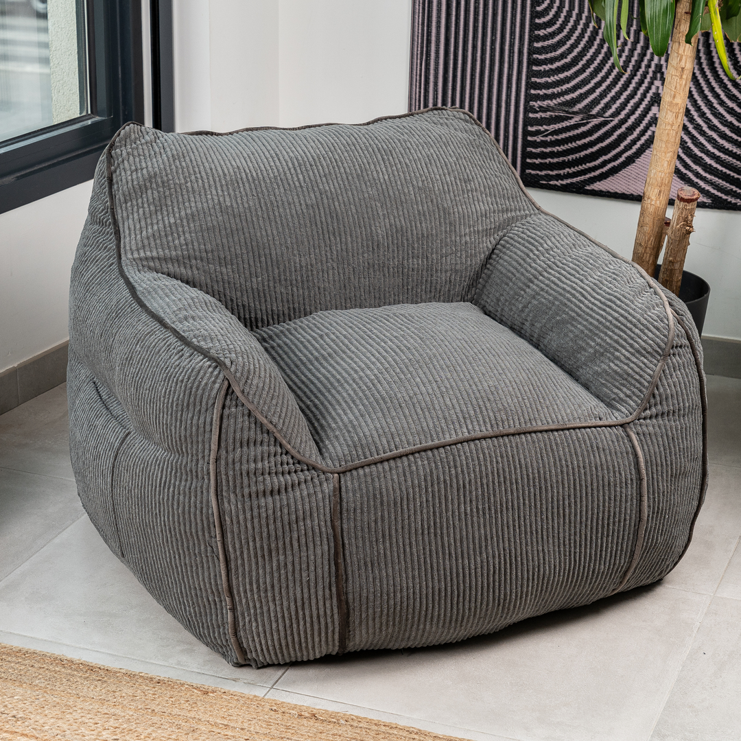 Pouf fauteuil velours côtelé gris foncé, forme compacte, accoudoirs intégrés, dans un salon